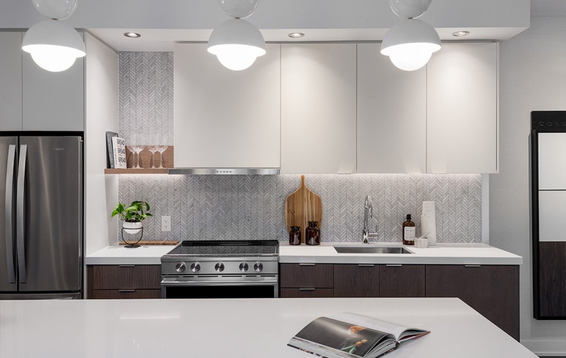 A modern kitchen with white cabinets, stainless steel appliances, and a patterned backsplash. Featuring a potted plant and cutting boards on the counter. Three hanging lights beautifully illuminate the space, perfect for new homes in Toronto. An open magazine rests on the island countertop.