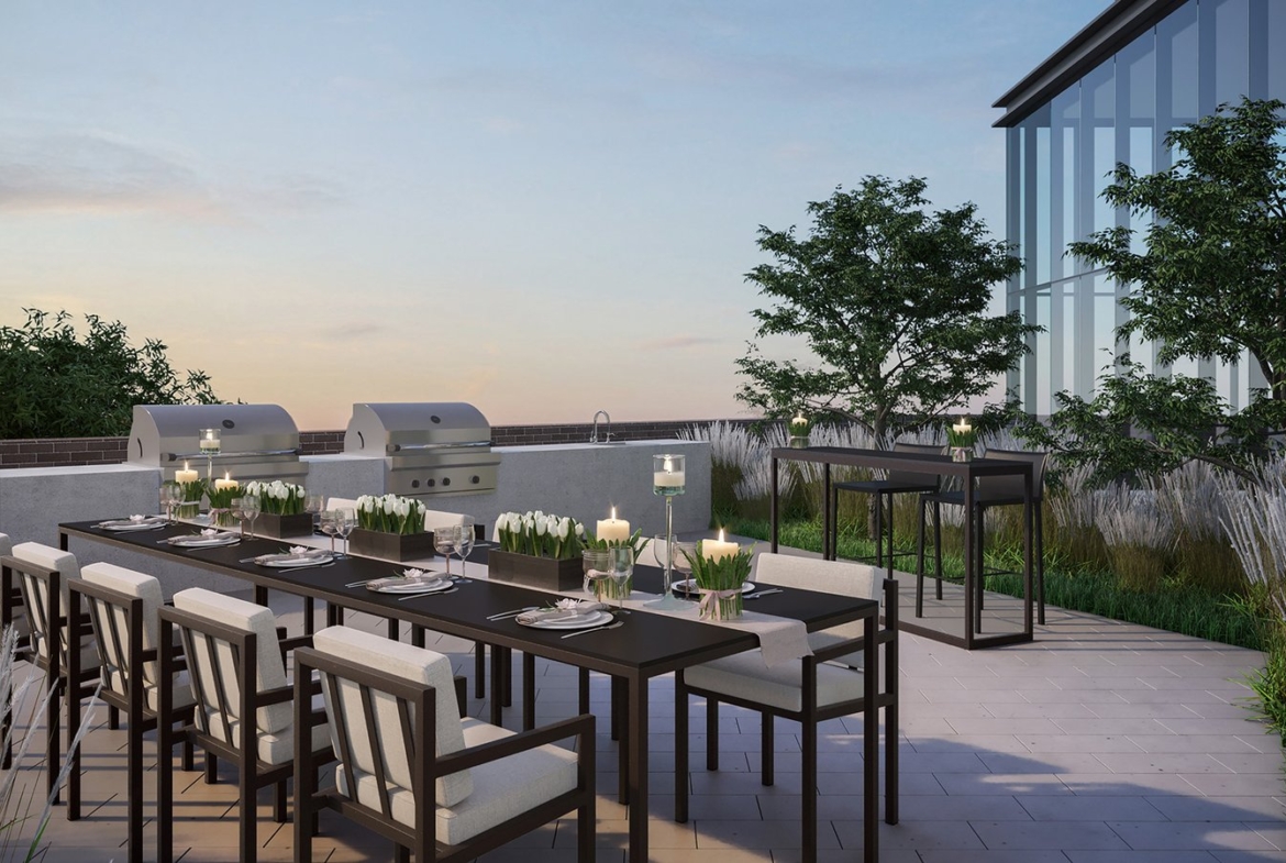 A modern outdoor patio in the new condos in Brampton features a long dining table with chairs, set for dinner with plates and candles. Two grills are on the side, and lush plants provide a scenic backdrop. The clear sky completes this serene evening setting.