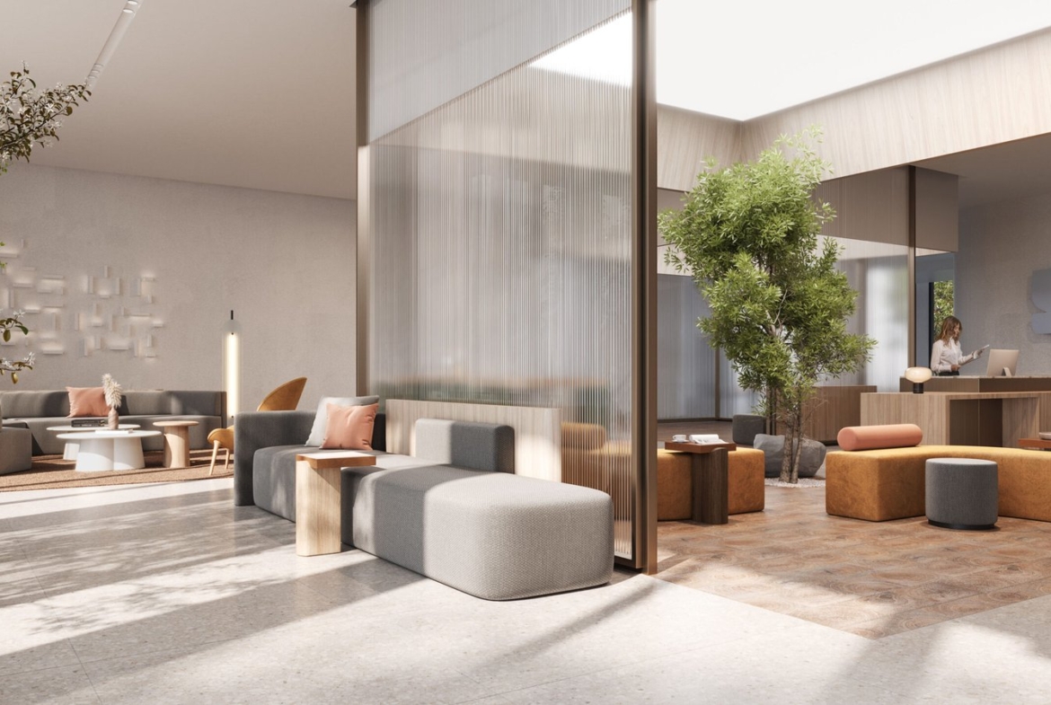 In this sophisticated condominium lobby, sleek gray and brown furniture, including sofas and ottomans, complement the space. A tree and plants add greenery near a wooden reception desk where a person stands. Soft natural light filters through large windows, enhancing the welcoming atmosphere.