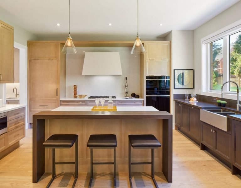 A modern kitchen in new homes features wooden cabinetry, a large island with three stools, pendant lights, and a built-in oven. A window above the double sink streams natural light into the room adorned in neutral tones.