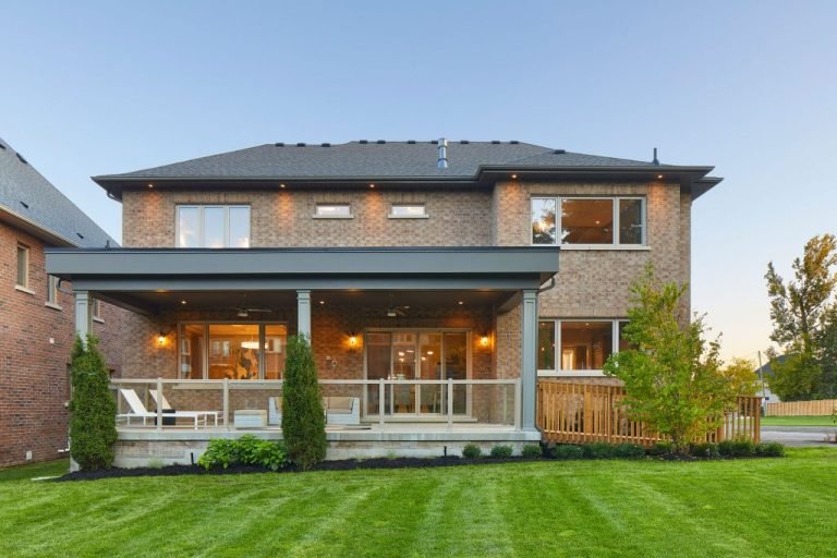 A charming two-story brick house with a wide wooden porch and pillars, surrounded by a well-maintained lawn and shrubs. Large windows grace both floors, basking in sunlight. It's the ideal inspiration for those considering new homes in GTA.