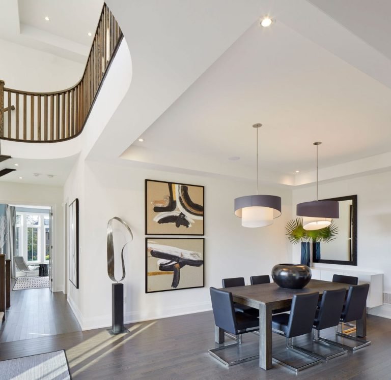 In a modern dining room typical of new homes in GTA, a dark wood table and six black chairs form the centerpiece. Two large abstract paintings adorn the white walls, complemented by pendant lights overhead. A unique sculpture graces the curved staircase railing as natural light floods the space.