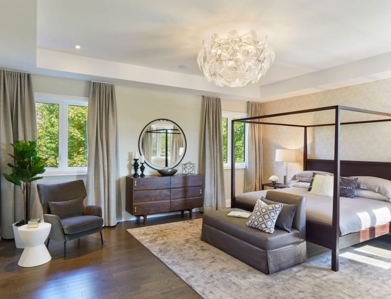 A modern bedroom in one of the new homes features a canopy bed, patterned pillows, and a bench at the foot. A dresser with a round mirror, an armchair, and a side table accent the space. Large windows with beige curtains and a statement chandelier complete the room's airy elegance.