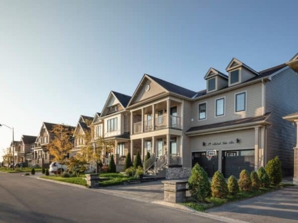 A row of modern suburban houses, including new homes in GTA, with well-maintained lawns and driveways, viewed during daytime under a clear blue sky. Some homes feature front porches and balconies, with small trees and shrubs lining the street.
