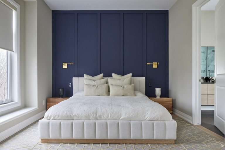 A modern bedroom in new condos features a large bed with white pillows and a cushioned headboard. The deep blue wall with gold accents enhances elegance, while natural light from the window highlights the room's luxurious feel. A glimpse of the bathroom completes this Gagan Bector masterpiece.