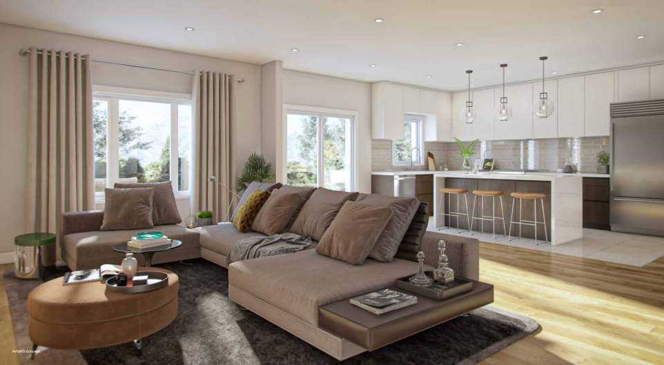 A modern, open-plan living room and kitchen in Sienna Woods. Features a large beige sectional sofa with brown pillows, a round coffee table, and a kitchen island with bar stools. Sunlight streams through large windows, highlighting the wooden floors and the charm of this tranquil town.