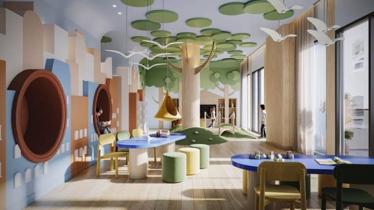 A brightly lit children's playroom with a nature theme, perfect for families in Spadina condos. The walls boast murals of trees, and the ceiling has hanging green circles simulating foliage. Play areas feature tunnels, tables, and chairs, while large windows let in natural light at 101 Spadina.