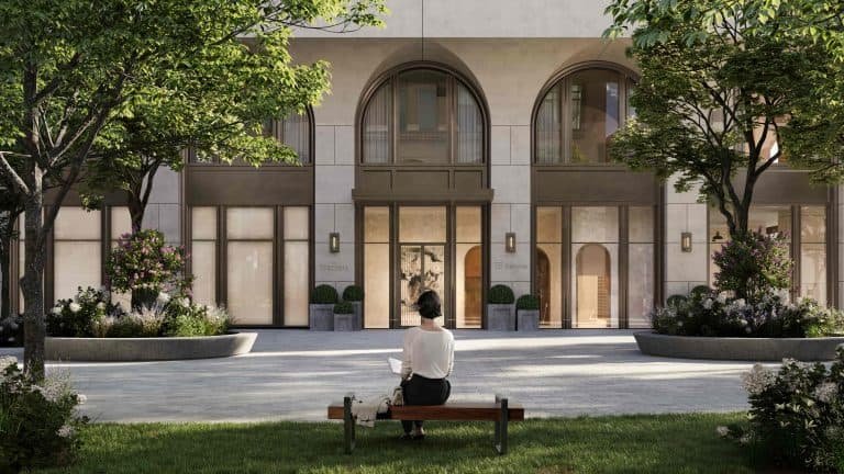 A person sits on a bench reading a book in front of the 101 Spadina Condos, a modern building with large arched windows and a central entrance. The surrounding area features trees and greenery, contributing to a serene, park-like setting. The weather appears clear and sunny.