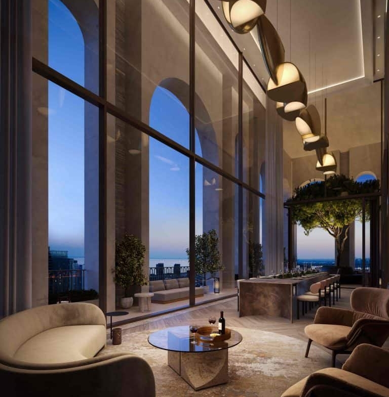 A luxurious, modern lounge at 101 Spadina Condos with floor-to-ceiling windows showcasing a cityscape at dusk. The room features elegant, curved sofas, a glass coffee table with a wine bottle and glasses, plants, and a sleek, suspended light fixture. Large arches frame the windows.