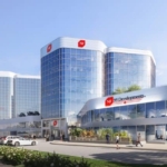 A modern office building with a reflective glass facade, labeled "NF Developments," stands amidst lush greenery and a bustling parking lot. It mirrors the ambitions of Gagan Bector in pre-construction projects. Above, a clear blue sky and distant airplane trail complete the scene.