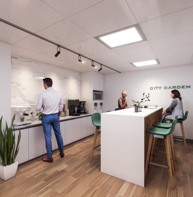 In the modern office kitchen of Gagan Bector, two women are seated at a tall table while a man in a white shirt and blue pants brews coffee. The space boasts white cabinets, wooden flooring, a plant, and the words "CITY GARDEN" elegantly displayed on the wall.