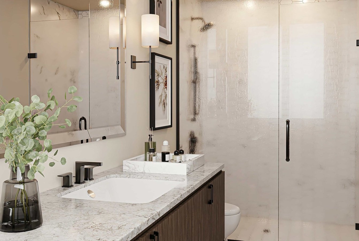 Modern bathroom in new GTA condos featuring a marble countertop with a sleek sink, a vase with greenery, and various toiletries. A large glass shower enclosure with a black handle is in the background, alongside framed artwork on the walls.