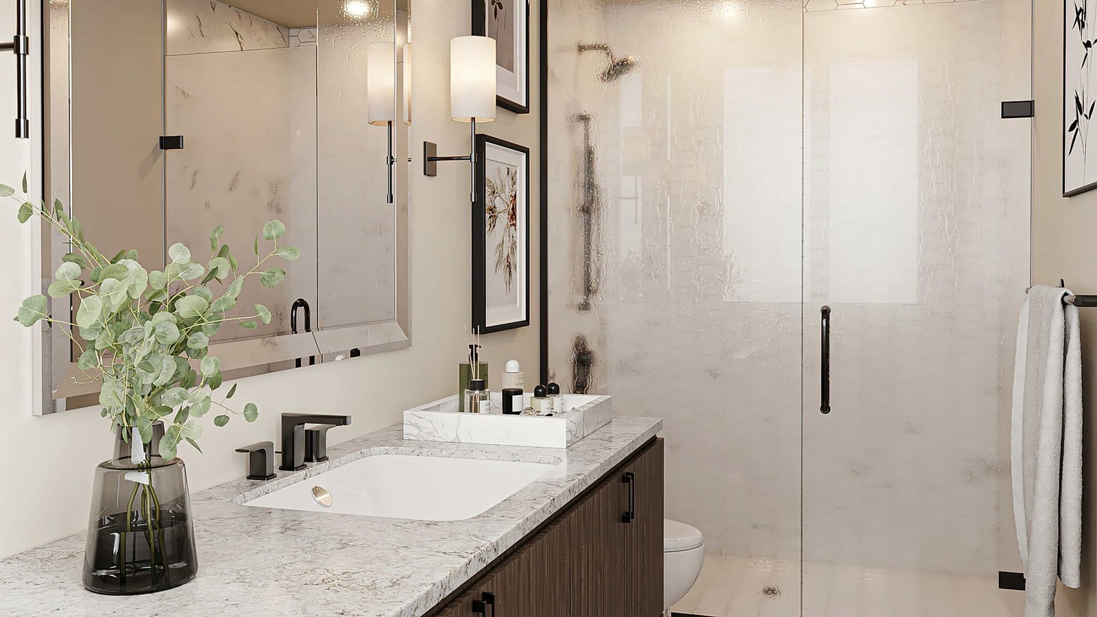 Modern bathroom in new GTA condos featuring a marble countertop with a sleek sink, a vase with greenery, and various toiletries. A large glass shower enclosure with a black handle is in the background, alongside framed artwork on the walls.