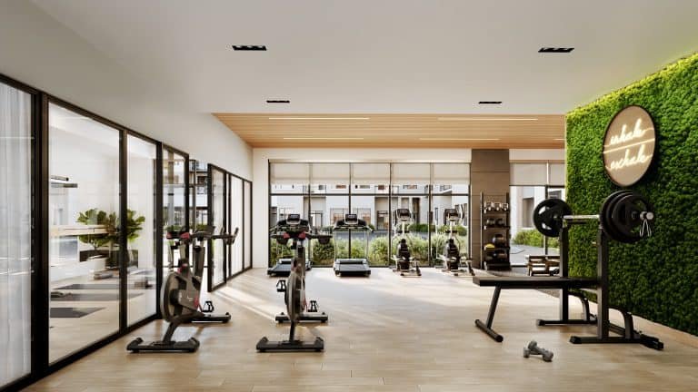 A modern gym in new condos features wooden flooring, stationary bikes, weightlifting equipment, and a treadmill. A green wall with a neon sign adds a fresh touch. Large windows let in natural light, illuminating the space and highlighting the luxury of pre-construction homes.
