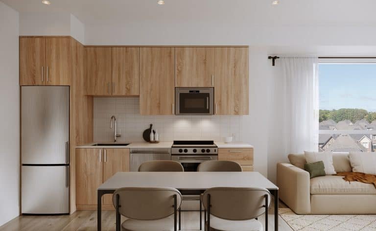 A modern kitchen in new homes in GTA features light wood cabinets, stainless steel appliances, and a white countertop. A dining table with four chairs sits nearby. To the right, a cozy living area with a beige sofa offers a bright window view, perfect for unwinding.