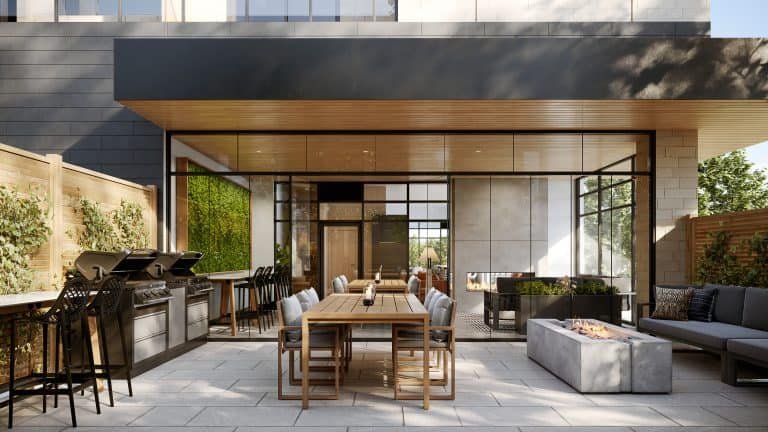 Modern outdoor patio space in new GTA homes features a long wooden dining table with chairs, a built-in grill set, and a rectangular fire pit. The patio is enclosed by a wooden fence and greenery, with large glass doors leading into the building.
