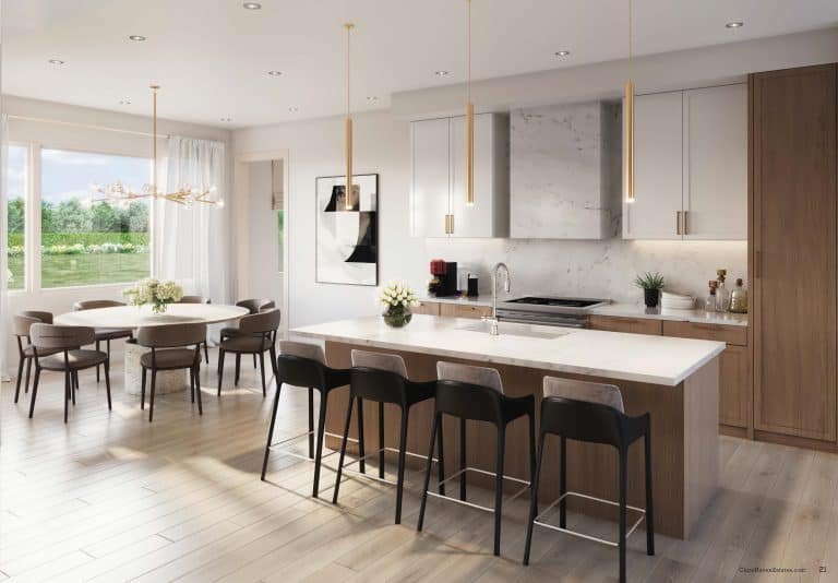 A modern, light-filled kitchen at Clarehaven Estates features a large island with wooden cabinets, white countertops, and four black barstools. Pendant lights hang above the island. A dining table with six chairs sits to the left, near sliding doors that open to a garden view.