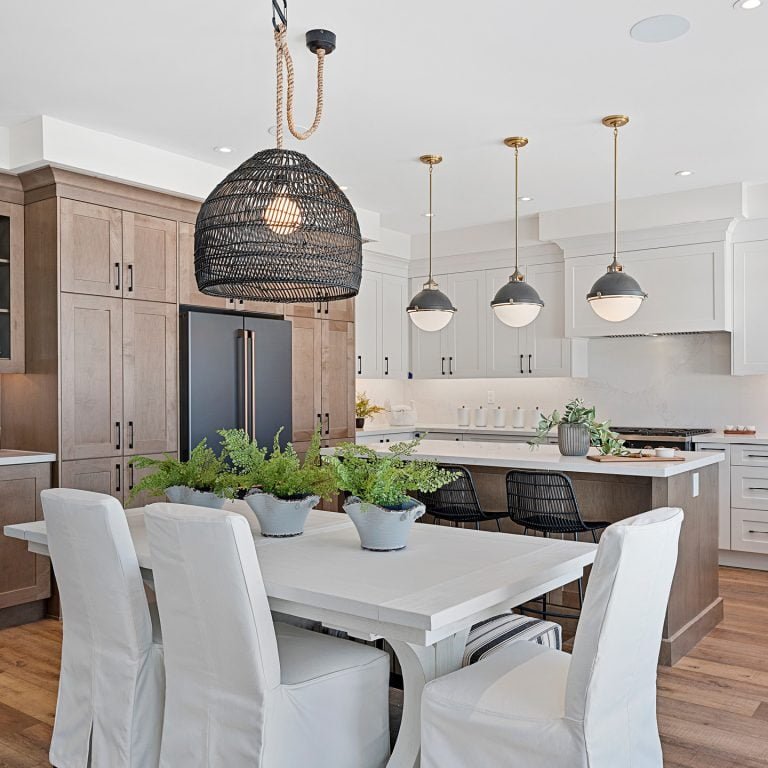Clarehaven Estates presents a modern kitchen and dining area featuring light wood cabinetry, a white island with black seating, and three pendant lights hanging above. A woven black chandelier is suspended over a white dining table with cushioned chairs and potted plants as a centerpiece.