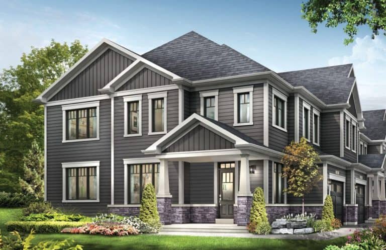 A two-story house with gray siding, white trim, and a dark gray roof stands proudly like a legacy of craftsmanship. The front entrance has a small covered porch. The yard is landscaped with green grass, colorful flowers, small bushes, and a tree. The sky above is clear and blue.