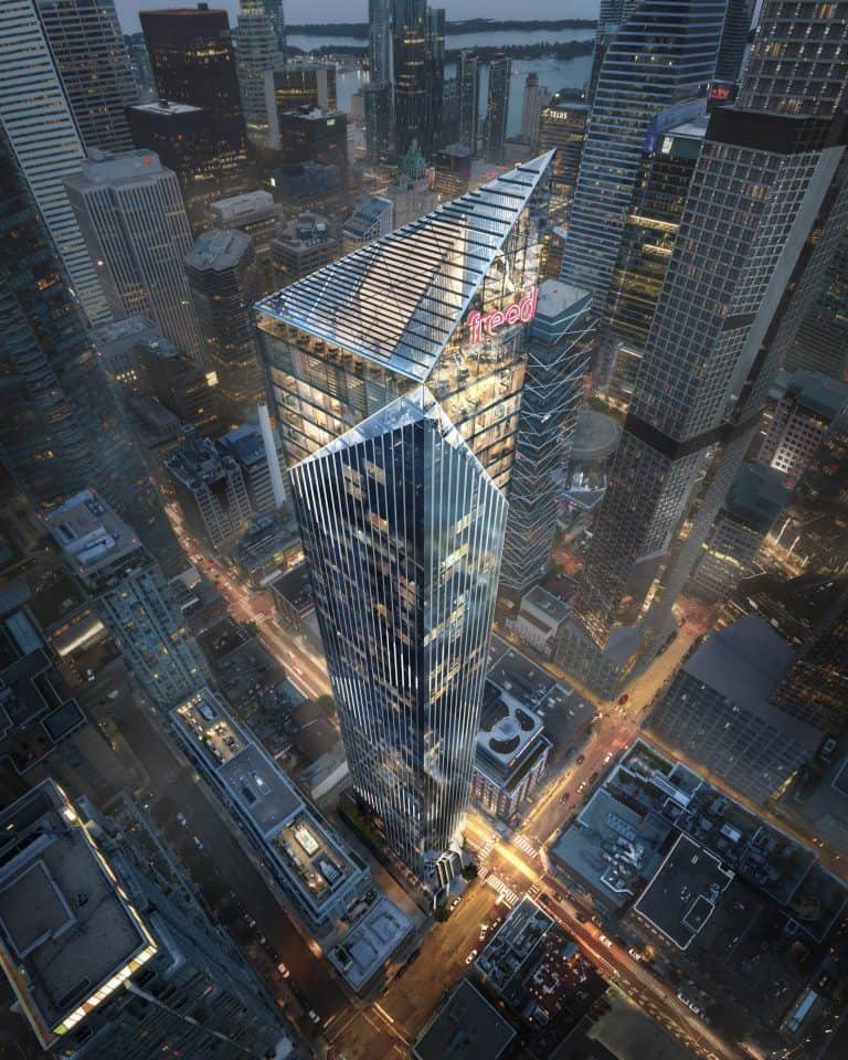 Aerial view of a modern cityscape featuring a tall, geometric skyscraper with a distinct angular design. The building is illuminated against the backdrop of neighboring high-rises and city streets lined with vehicles. Near the top, the sign reading "Freed Hotel" is visible among sleek residences.