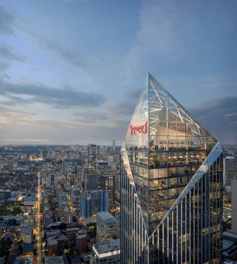 An aerial view of a skyscraper with a distinctive illuminated triangular glass top, prominently displaying the red lettering "free'd". The cityscape below features various buildings and residences, with a street bustling with traffic, extending into the twilight horizon.