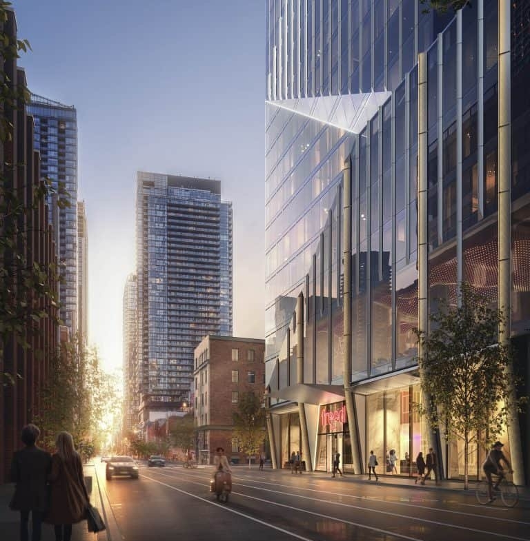 A bustling urban street at dusk, with modern high-rise buildings lining both sides. People walk along the sidewalks and a few cars drive down the road. The street is illuminated by city lights, including those from the nearby Freed Hotel Residences, and the setting sun in the background creates a warm glow.