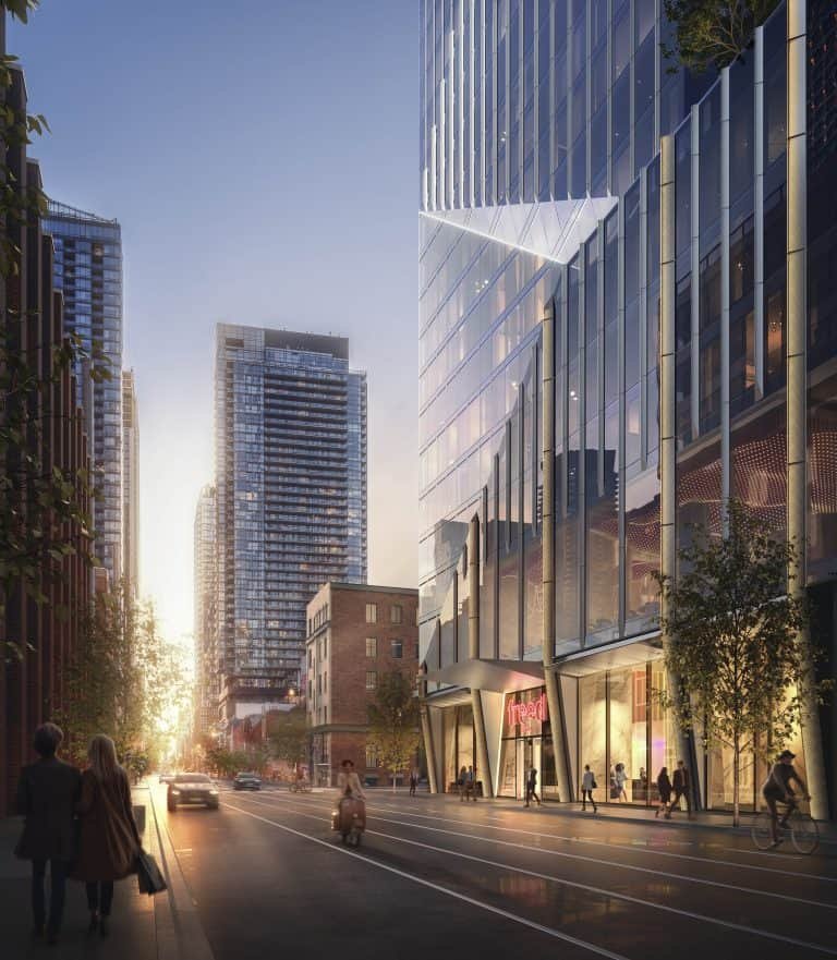 A bustling urban street at dusk, with modern high-rise buildings lining both sides. People walk along the sidewalks and a few cars drive down the road. The street is illuminated by city lights, including those from the nearby Freed Hotel Residences, and the setting sun in the background creates a warm glow.