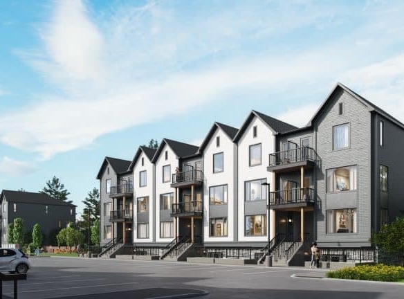 A row of modern, three-story townhouses designed by Gagan Bector stands with gabled roofs and large windows. The buildings, painted in neutral colors, overlook a mostly empty parking lot with a few cars. Under the clear blue sky, these pre-construction homes exude contemporary charm.