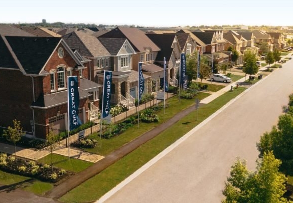 Aerial view of a residential neighborhood featuring pre-construction homes with modern two-story designs. Each house boasts a front yard and driveway. Tall banners line the tree-lined street, running parallel to these new homes in GTA, all under a clear sky.