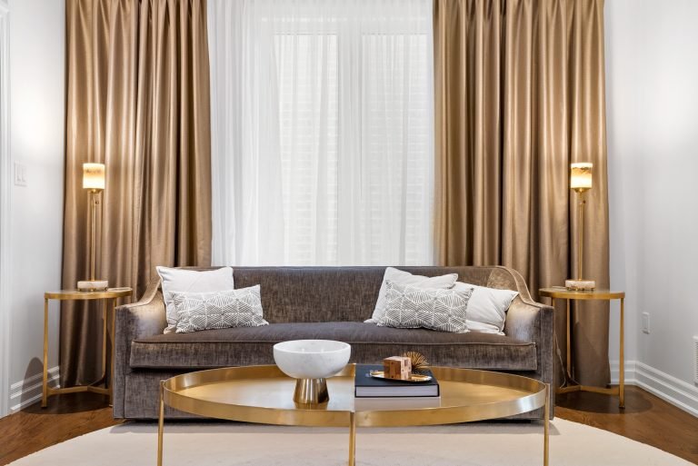 A modern living room by Gagan Bector features a gray couch adorned with white and patterned pillows. Two gold side tables with lamps complement a large round coffee table. Beige curtains frame sheer white drapes, enhancing the elegant feel of these pre-construction homes.