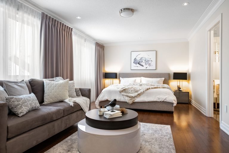 A modern bedroom in one of the new condos features a large bed with white bedding and a faux fur throw. A beige sofa with cushions sits in the foreground, while a round coffee table holds decorative items. Two side tables with black lamps flank the bed, and natural light floods the inviting space.