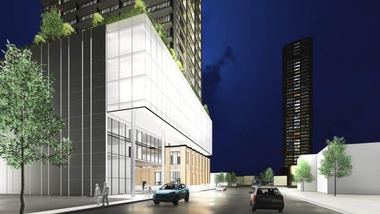 A nighttime view of a modern city street showcases tall buildings in the foreground. A glass-paneled structure with rooftop plants hints at new condos, as two cars glide along the street. Pedestrians stroll nearby, while another towering building stands proudly in the background.