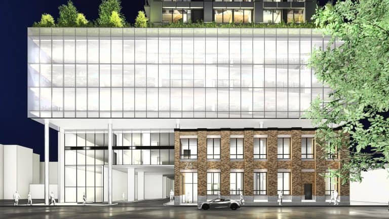 Modern building at night blending glass and brick architecture. The lower section features brick with tall windows, while the sleek, glass-fronted upper part exudes elegance. A few trees adorn the rooftop of these new homes in GTA. A car is parked in front as people stroll by.