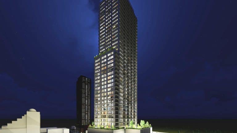 Night view of a tall, illuminated skyscraper with numerous lit windows surrounded by smaller buildings. The clear, dark sky enhances the building's lights, while new condos rise alongside. Trees frame the base of this modern marvel, offering a glimpse into urban living at its finest.