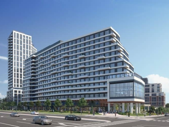 A modern urban intersection showcases a multi-story building with sleek glass and white facades. The scene, reminiscent of new condos' rising skyline, includes several cars on the street and a clear blue sky. People stroll past the building, where trees line the sidewalks gracefully.