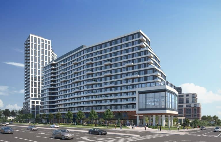 A modern urban intersection showcases a multi-story building with sleek glass and white facades. The scene, reminiscent of new condos' rising skyline, includes several cars on the street and a clear blue sky. People stroll past the building, where trees line the sidewalks gracefully.