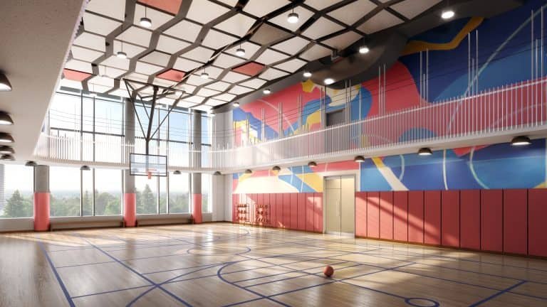 A modern indoor basketball court in Gagan Bector features a wooden floor and a colorful geometric mural on the walls. Sunlight streams through large windows, highlighting the red padded walls. A basketball rests on the court as ceiling lights illuminate this perfect addition to new homes in GTA.
