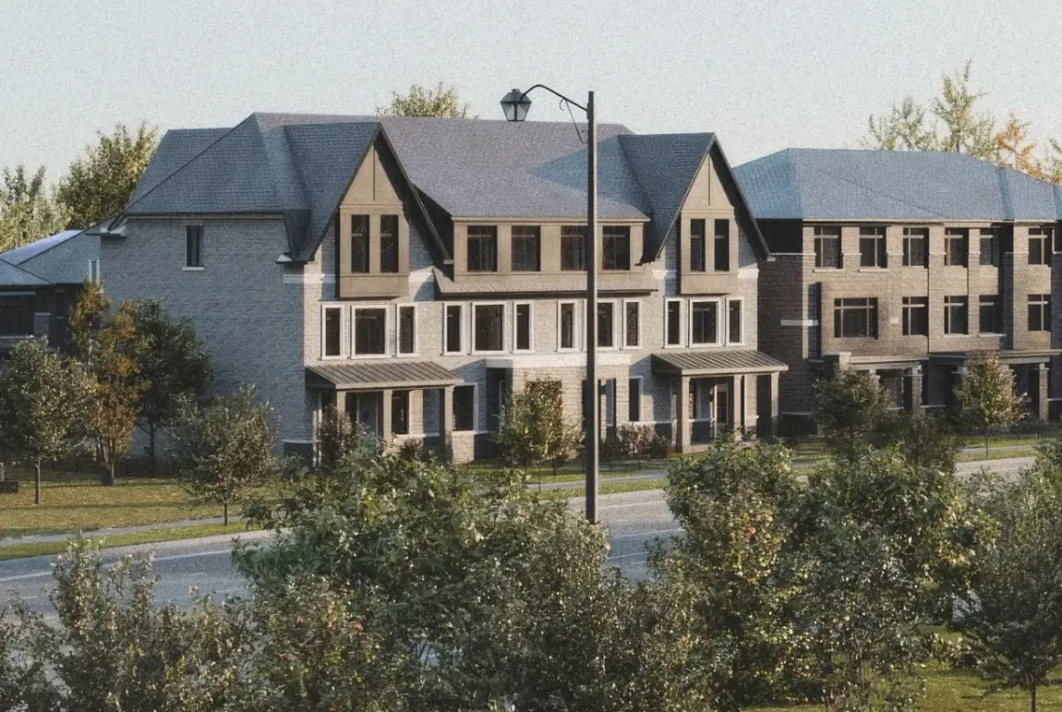 A row of modern townhouses with sloped roofs is situated behind a street lined with trees and grass. These new homes offer a fresh take on urban living. In the foreground, there is a small parking area and a few traffic cones. The sky is clear.