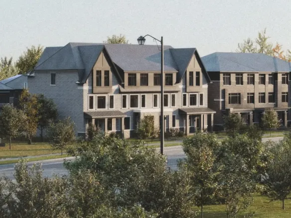 A row of modern townhouses with sloped roofs is situated behind a street lined with trees and grass. These new homes offer a fresh take on urban living. In the foreground, there is a small parking area and a few traffic cones. The sky is clear.