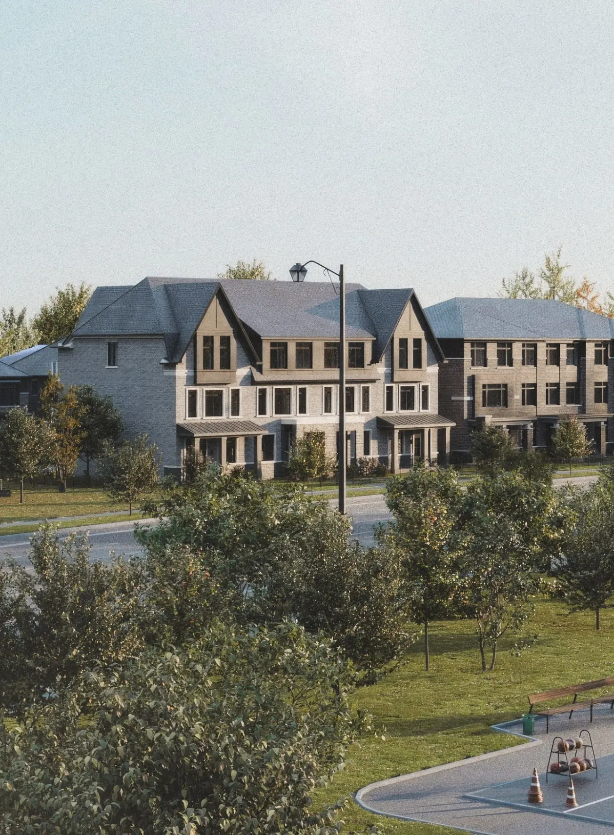 A row of modern townhouses with sloped roofs is situated behind a street lined with trees and grass. These new homes offer a fresh take on urban living. In the foreground, there is a small parking area and a few traffic cones. The sky is clear.