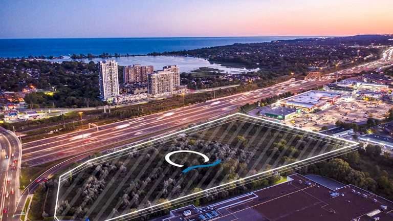 Aerial view of a cityscape at dusk, featuring a highway with mesmerizing light trails, surrounding buildings, and pre-construction homes outlined by white lines. In the background, there's a large body of water and new condos in the GTA among residential high-rises.