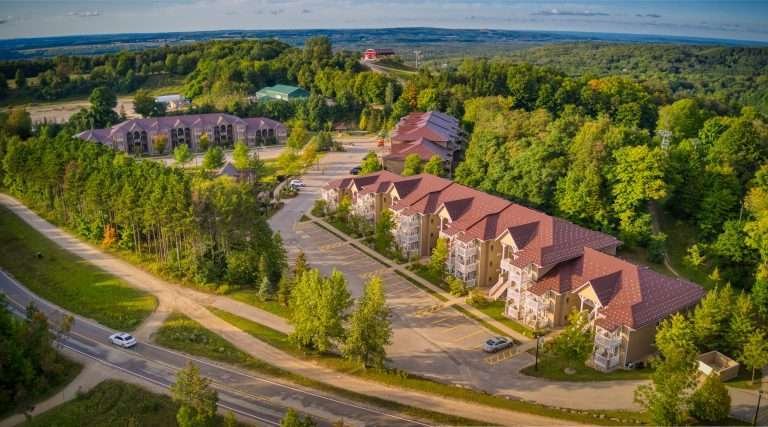 Aerial view of a residential area featuring rows of large, multi-story buildings with reddish-brown roofs, surrounded by lush green trees, near the Carriage Country Club, under a clear sky.