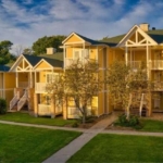 Aerial view of the Carriage Country Club apartment complex featuring multiple three-story buildings with balconies and external staircases, surrounded by lush greenery and a well-maintained lawn, bathed in warm sunlight.