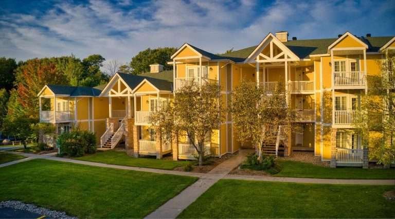 Aerial view of the Carriage Country Club apartment complex featuring multiple three-story buildings with balconies and external staircases, surrounded by lush greenery and a well-maintained lawn, bathed in warm sunlight.